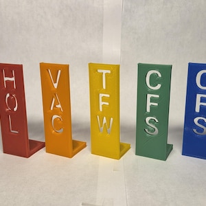 May include: Five colorful, 3D-printed signs in red, orange, yellow, green, and blue. Each sign has a different three-letter word cut out: HOL, VAC, TFW, CFS, and CFS. The signs are standing upright.