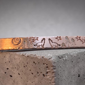 May include: Copper-colored bracelet with etched design. Features a crescent moon and star, pine trees, and two howling wolves under stars. The bracelet is a unique piece of jewelry.