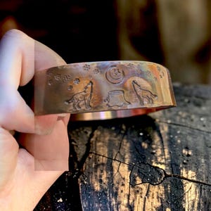 May include: Copper cuff bracelet with an engraved wolf howling at the moon design. The bracelet features two wolves, a crescent moon, stars, and a tree stump. The metal has a warm, reddish-brown hue.