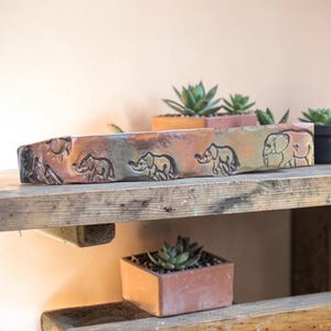 May include: A rectangular, handmade planter with a textured, metallic finish. The planter features embossed elephant designs and a sun and mountain scene. The planter is displayed on a rustic wooden shelf, with other small potted succulents.
