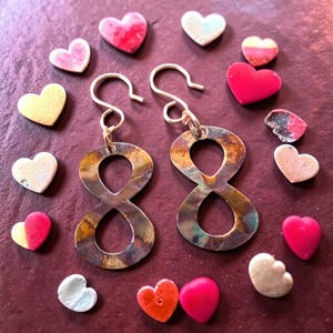 May include: Artisan earrings in the shape of the number eight, with a rustic, multi-toned finish. The earrings are displayed with an assortment of heart-shaped beads in various colors, including pink, red, and white. The earrings have silver-colored ear wires.