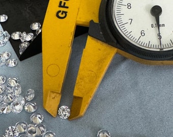 Pode incluir: Close-up de um paquímetro GFC amarelo medindo um pequeno diamante transparente. O diamante é cercado por uma dispersão de outros diamantes em uma superfície de tecido azul claro.