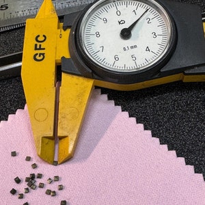 May include: A yellow GFC caliper measuring a pile of small, dark gray, square-shaped metal pieces on a pink fabric surface.