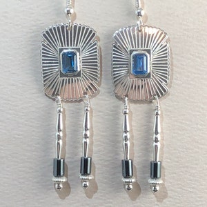 May include: Silver dangle earrings with a blue gemstone set in a square frame and three black beads hanging from each earring.
