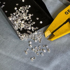 May include: A close-up of a black tray filled with loose diamonds. The diamonds have spilled out onto a gray surface. A yellow tool with the word "GE" printed on it is lying on the surface.