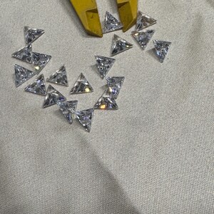 May include: A collection of small, clear, triangular-shaped gemstones. The gemstones are scattered on a light-colored surface, with a yellow tool partially visible at the top. The gemstones appear to be cut with multiple facets.