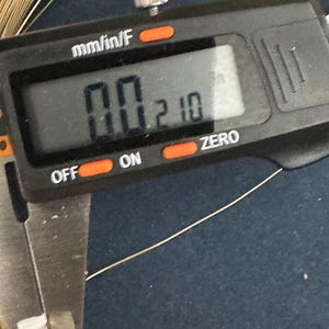 May include: A close-up of a digital caliper measuring a thin gold wire. The caliper reads "0.0210" mm/in/F.