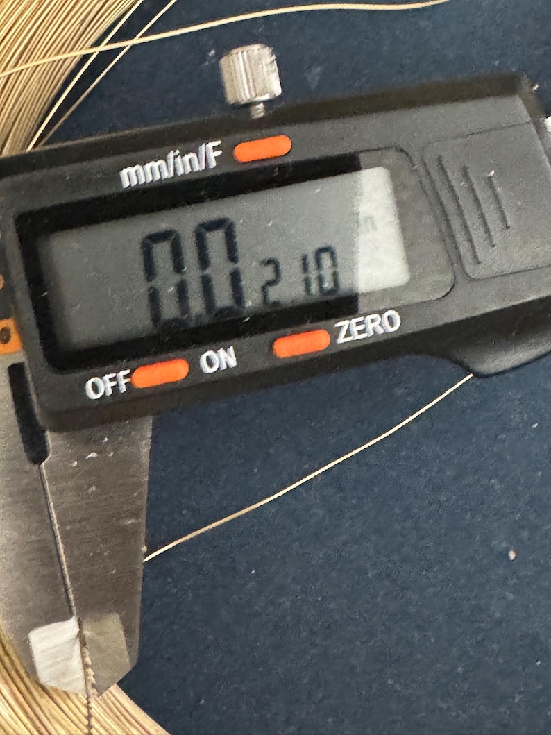 May include: A close-up of a digital caliper measuring a thin gold wire. The caliper reads "0.0210" mm/in/F.