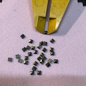 Könnte beinhalten: Nahaufnahme einer Gruppe kleiner, schwarzer, pyramidenförmiger Strasssteine, die auf einer rosa Stoffoberfläche verstreut sind. Die Strasssteine sind in einem zufälligen Muster angeordnet.