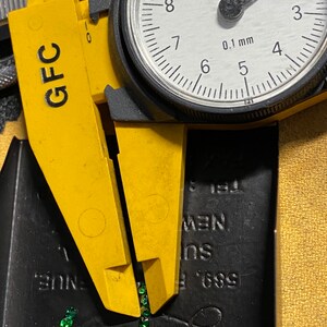 May include: A yellow caliper with a dial gauge measuring in millimeters. The caliper is open and there are small green gemstones scattered on a black surface.