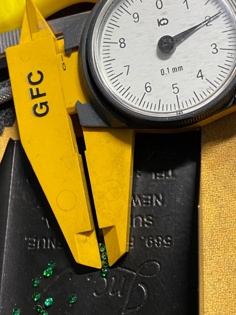 May include: A yellow caliper with a dial gauge measuring in millimeters. The caliper is open and there are small green gemstones scattered on a black surface.