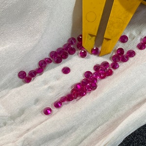 May include: A collection of small, round, faceted, deep pink gemstones scattered on a white surface. A yellow caliper is partially visible, measuring the gems. The gems are likely rubies, and the image suggests jewelry making or gem evaluation.
