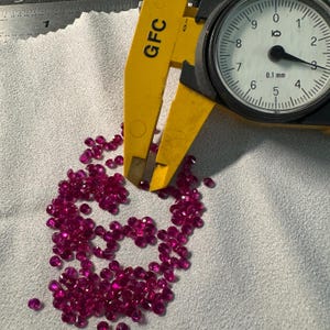 Op de afbeelding: Een close-up van een stapel kleine, felroze edelstenen die worden gemeten met een gele schuifmaat. De schuifmaat geeft 0,1 mm aan.