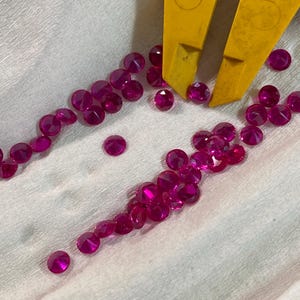 May include: A collection of loose pink gemstones, likely rubies, scattered on a white surface. Some of the gemstones are arranged in a line, while others are scattered randomly.
