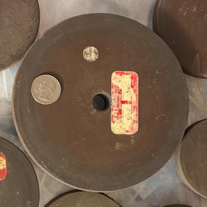 May include: A collection of brown, circular grinding wheels with a central hole. One wheel has a quarter coin next to it for scale. A red and white sticker with text is affixed to one wheel.