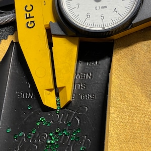 May include: A yellow caliper with the text "GFC" on it is measuring a pile of small, green, faceted gemstones. The caliper is set to 0.1 mm and the dial reads 6.8. The gemstones are scattered on a black surface with the text "589 F NUE, Ins NEW" printed on it.