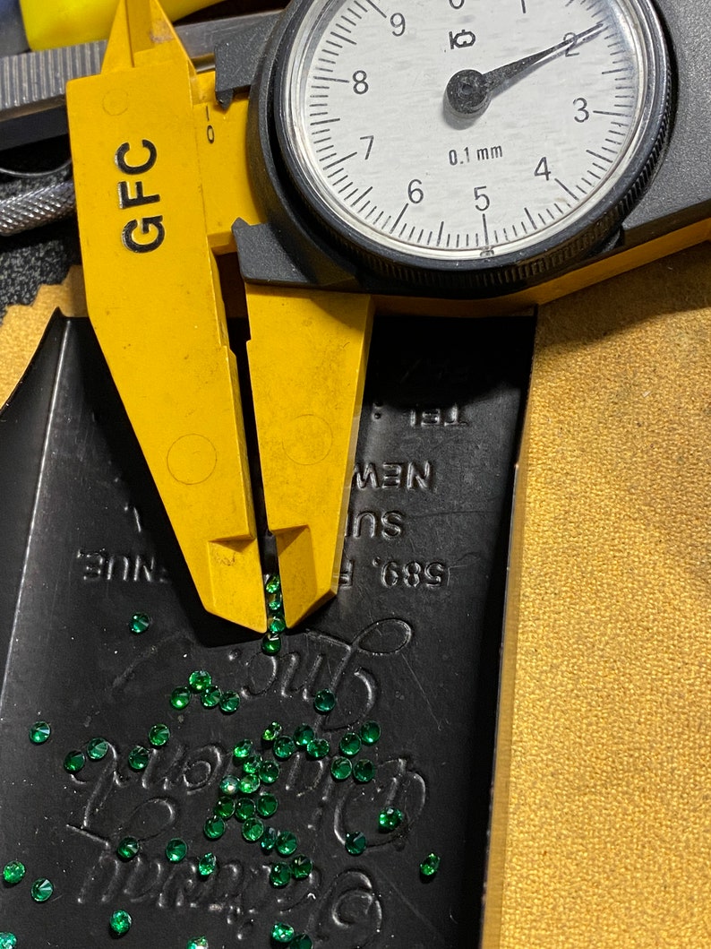 May include: A yellow caliper with the text "GFC" on it is measuring a pile of small, green, faceted gemstones. The caliper is set to 0.1 mm and the dial reads 6.8. The gemstones are scattered on a black surface with the text "589 F NUE, Ins NEW" printed on it.