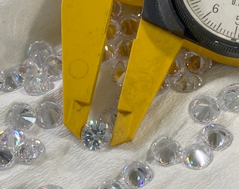 May include: A yellow caliper with the text "GFC" on it is used to measure a clear, faceted gemstone. The caliper has a dial gauge with markings from 5 to 9. The gemstone is surrounded by other clear, faceted gemstones on a white surface.