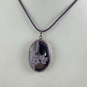 May include: A silver oval locket necklace with a purple cord. The locket has a black and white geometric design with pink flowers.