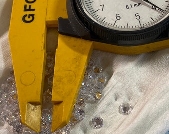 May include: A yellow caliper with the text "GFC" on the side, measuring a pile of small, clear, round gemstones. The caliper has a dial that reads "0.1 mm" and is set to 4.5 mm.