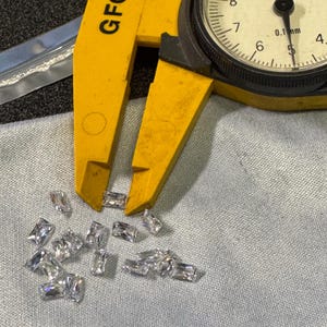 Op de afbeelding: Een gele schuifmaat met een wijzerplaat die 0,1 mm aangeeft, is open en rust op een grijs stofoppervlak. Een strooiing van heldere, rechthoekige edelstenen is verspreid over de stof.