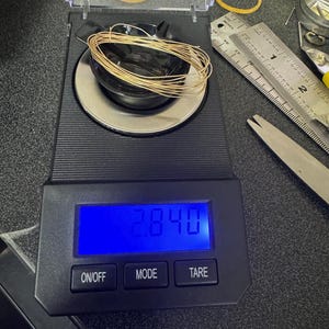 May include: A digital scale with a black base and a blue LCD screen displaying "28.40". The scale has three buttons labeled "ON/OFF", "MODE", and "TARE". A coil of gold wire is sitting on the scale.