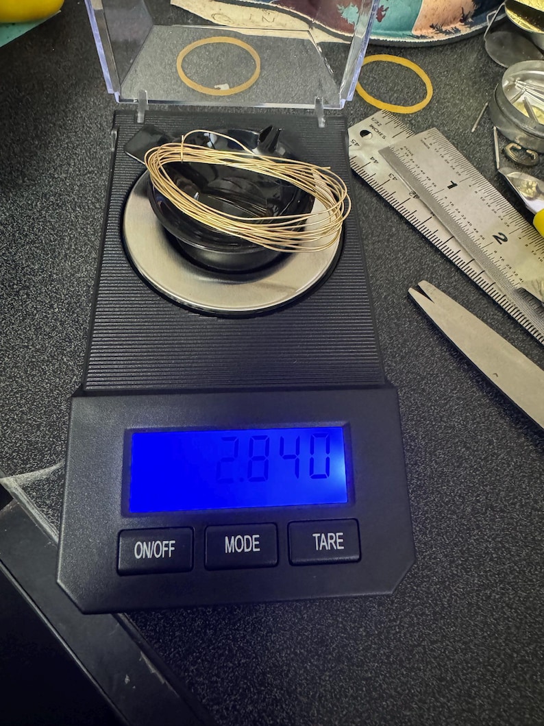 May include: A digital scale with a black base and a blue LCD screen displaying "28.40". The scale has three buttons labeled "ON/OFF", "MODE", and "TARE". A coil of gold wire is sitting on the scale.