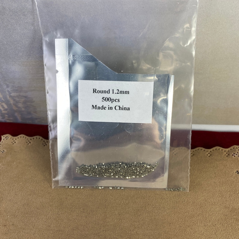 K&ouml;nnte beinhalten: Eine transparente Plastikt&uuml;te mit einem kleinen Haufen runder, silberfarbener Metallperlen. Die T&uuml;te tr&auml;gt ein Etikett mit der Aufschrift "Round 1.2mm 500pcs Made in China".