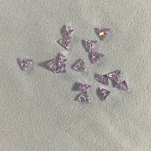 May include: A collection of small, triangular, clear, iridescent purple rhinestones. The rhinestones are scattered on a light gray fabric surface.