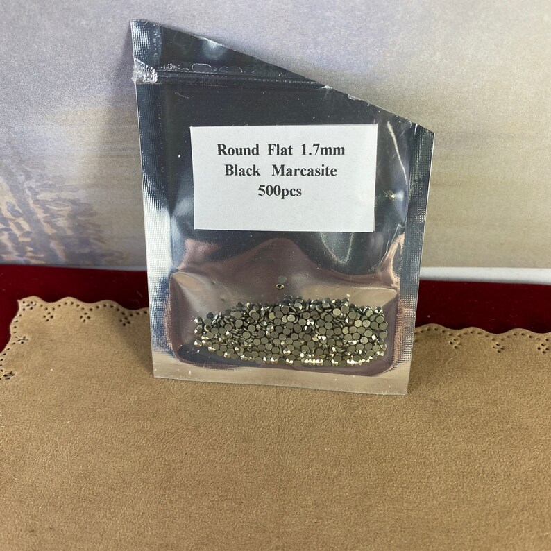 K&ouml;nnte beinhalten: Ein transparenter Plastikbeutel mit 500 kleinen, runden, flachen, schwarzen Markasit-Strasssteinen. Der Beutel tr&auml;gt ein Etikett mit der Aufschrift "Round Flat 1.7mm Black Marcasite 500pcs".