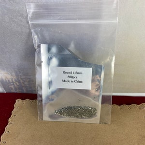 K&ouml;nnte beinhalten: Eine transparente Plastikt&uuml;te mit einer kleinen Menge silberfarbener runder Strasssteine. Die T&uuml;te tr&auml;gt ein Etikett mit der Aufschrift "Round 1.5mm 500pcs Made in China."
