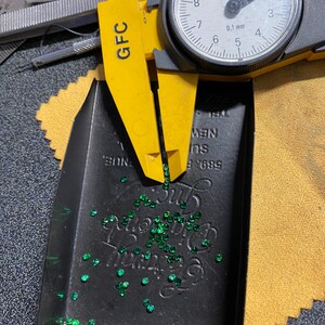 May include: A close-up of a black tray filled with small, green gemstones. A yellow caliper with a dial gauge is measuring the gemstones. The dial gauge reads 0.1 mm. The tray has the text "589 E. SUE NEW 191" printed on it.