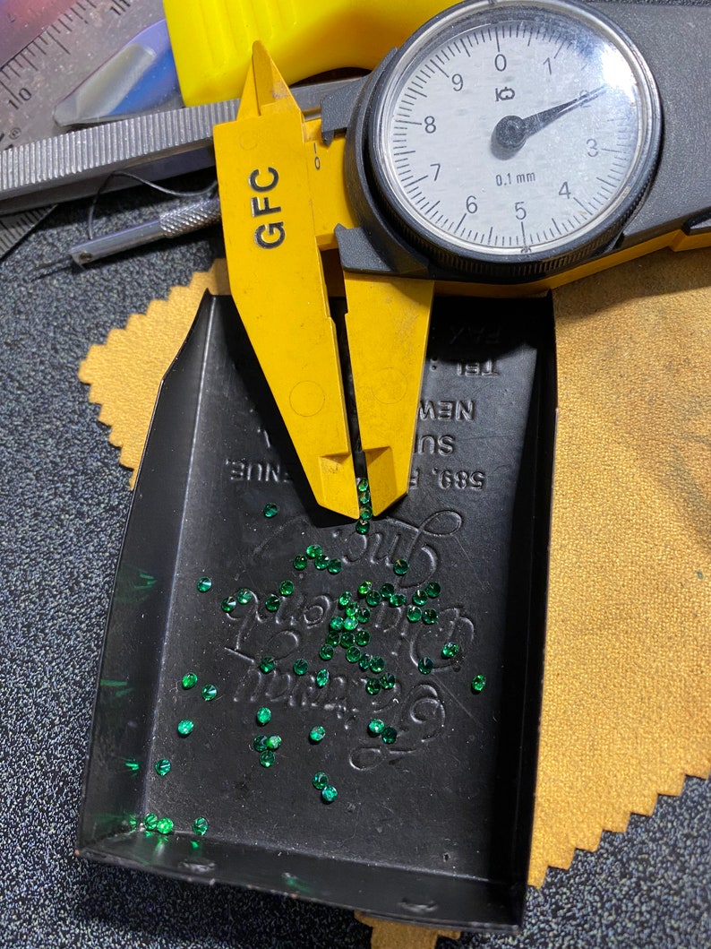 May include: A close-up of a black tray filled with small, green gemstones. A yellow caliper with a dial gauge is measuring the gemstones. The dial gauge reads 0.1 mm. The tray has the text "589 E. SUE NEW 191" printed on it.