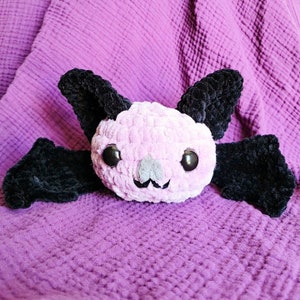 May include: A handmade plush bat with a light pink body, black wings and ears, and large, shiny black eyes. The bat has a gray nose and a smiling mouth. The plush toy is set against a purple background.