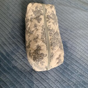 May include: A quilted toile-patterned zippered pouch with a white background and grey floral design.