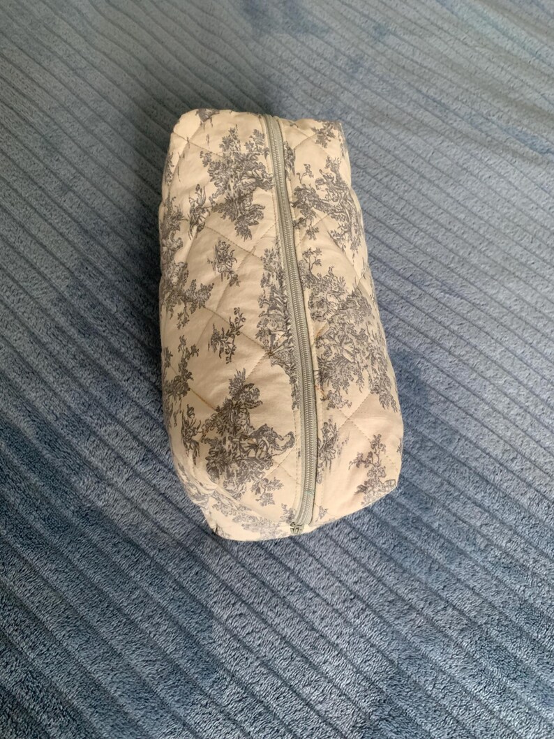 May include: A quilted toile-patterned zippered pouch with a white background and grey floral design.