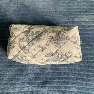 May include: A quilted toile-patterned makeup bag with a zipper closure. The bag is beige with a black and white toile pattern.