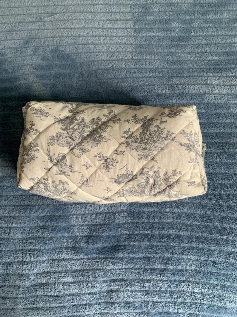 May include: A quilted toile-patterned makeup bag with a zipper closure. The bag is beige with a black and white toile pattern.