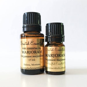 Puede incluir: Dos botellas de vidrio ámbar con tapas negras que contienen aceite esencial de mejorana. Las botellas tienen etiquetas doradas con el texto "Created Essentials" y "Pure Essential Oil Marjoram Origanum majorana 15 ml".