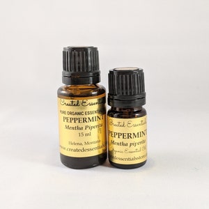 Puede incluir: Dos botellas de vidrio ámbar con tapas negras y etiquetas doradas. La botella más grande está etiquetada "Created Essentials Pure Organic Essential Oil Peppermint Mentha Piperita 15 ml Helena, Montana www.createdessentials.com". La botella más pequeña está etiquetada "Created Essentials Peppermint Mentha piperita Organic Essential Oil createdessentialsstore.com".