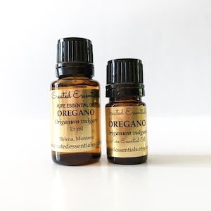 Op de afbeelding: Twee bruine glazen flessen oregano etherische olie. De etiketten lezen "Created Essentials" en "OREGANO" met de Latijnse naam "Origanum vulgare". De grotere fles bevat 15 ml olie. De kleinere fles is ook gelabeld met dezelfde informatie.