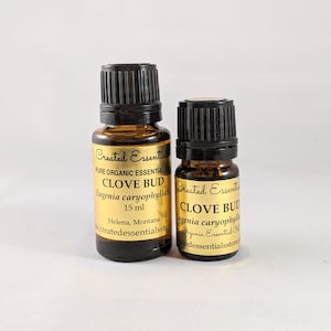 Może przedstawiać: Dwie bursztynowe butelki ze szklanymi korkami zawierające olejek eteryczny z goździków. Butelki są oznaczone etykietami "Created Essential" i "Clove Bud" z nazwą botaniczna "Eugenia caryophyllata". Większa butelka ma pojemność 15 ml, a mniejsza 10 ml.