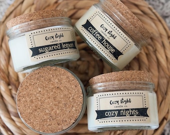 Puede incluir: Tres frascos de vidrio pequeños llenos de velas de cera blanca, cada uno con una tapa de corcho redonda. Las etiquetas muestran el texto Cozy Light candle co. con los nombres de aromas: sugared lemon, coffee house y cozy nights.