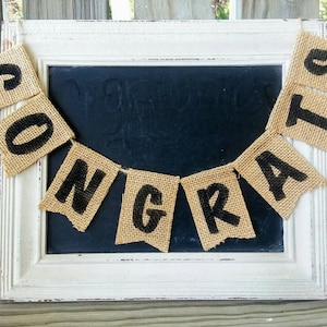 May include: A burlap banner with the word "Congrats" spelled out in black letters. The banner is hanging in front of a white framed chalkboard.