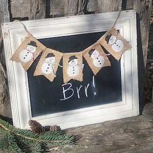 May include: A burlap banner with a string of snowmen hanging on a white framed chalkboard with the word "Brr!" written in white chalk.
