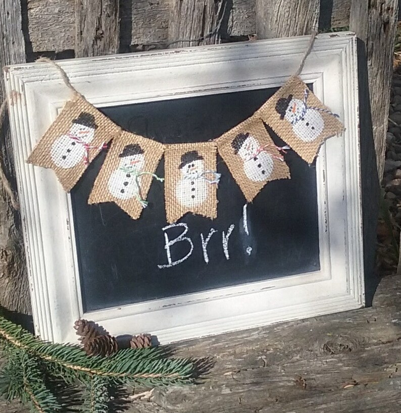 May include: A burlap banner with a string of snowmen hanging on a white framed chalkboard with the word "Brr!" written in white chalk.