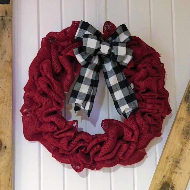 May include: A red burlap wreath with a black and white checkered bow. The wreath is made of burlap and has a ruffled texture. The bow is tied in a knot and has a long tail.