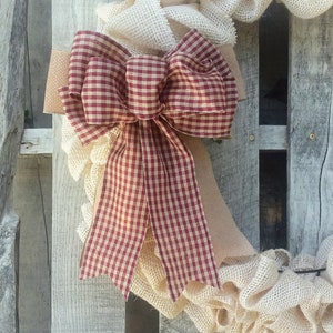 May include: A burlap wreath with a large red and white gingham bow. The wreath is made of burlap and has a rustic look.