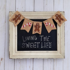 May include: A white framed chalkboard with the text "Living the Sweet Life" written in white chalk. A burlap banner with gingerbread men and candy canes hangs above the chalkboard.