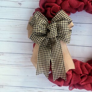May include: A red burlap wreath with a large black and tan gingham bow. The wreath is on a white wood background.